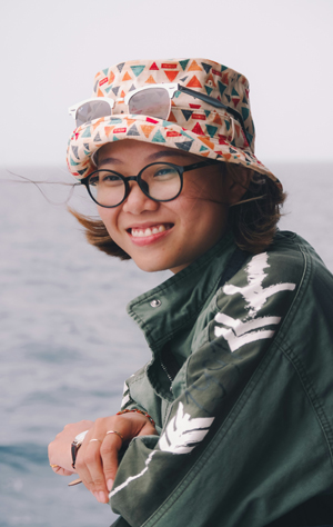 Une femme souriante d'origine asiatique, coiffée d'un chapeau à godets, est appuyée sur le rebord d'un bateau