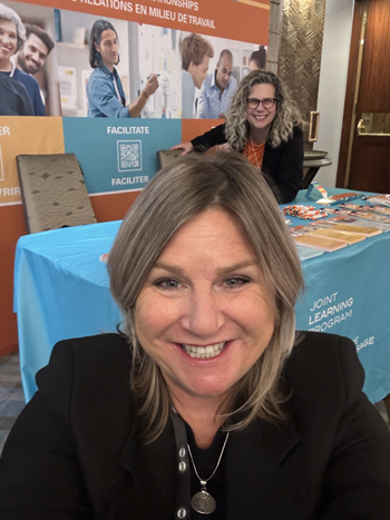 Catherine’s smiling face is close to the camera, while Roxanne poses behind her at the JLP kiosk.