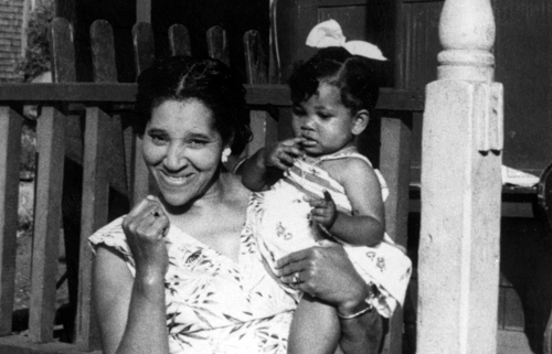 A young Black woman smiles for the camera. She is standing in front of a home and holding a small Black child in her arms. The picture is black and white, and was taken in Africville.