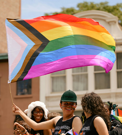 Deux personnes dans une foule se sourient. L&rsquo;une d&rsquo;elles brandit le drapeau de la Fiert&eacute; progressiste.