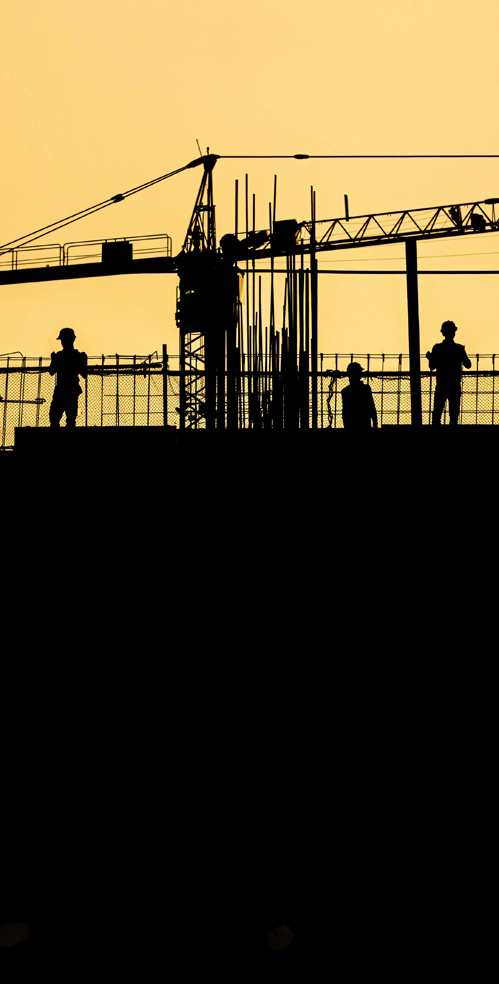 Workers in hardhats and construction rigging are silhouetted against a dramatic