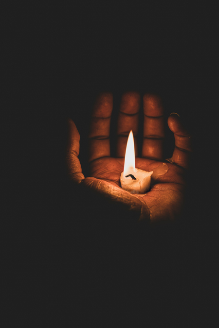 A close-up of a dark-skinned hand cupping a short lit candle