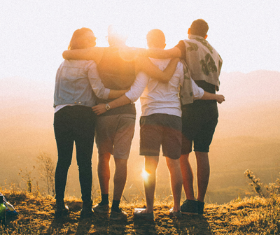 Four people stand in a line, admiring a sunset. Their backs are to the camera and they are physically supporting each other.