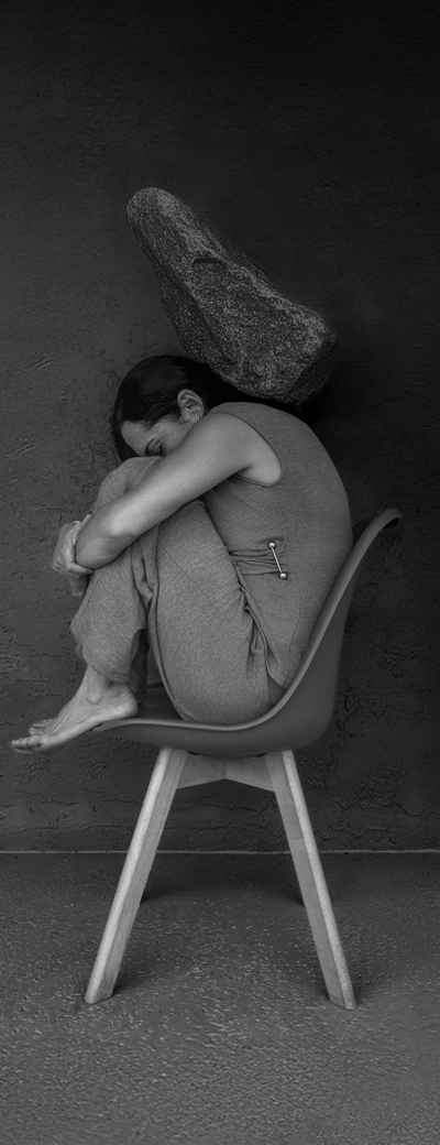 A black and white image of a woman seated on a stool with her arms wrapped around her legs.